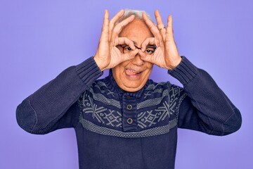 Canvas Print - Middle age senior grey-haired man wearing glasses and winter sweater over purple background doing ok gesture like binoculars sticking tongue out, eyes looking through fingers. Crazy expression.