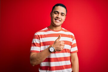 Wall Mural - Young brazilian man wearing casual striped t-shirt standing over isolated red background doing happy thumbs up gesture with hand. Approving expression looking at the camera showing success.