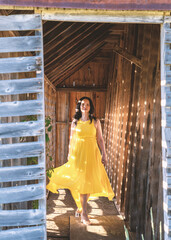 A close portrait of an Asian woman with long hair and yellow dress in front of an old barn on a farmland