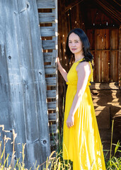 A close portrait of an Asian woman with long hair and yellow dress in front of an old barn on a farmland