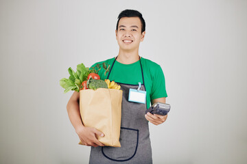 Portrait of smiling young supermarket worker with paper package with goceries and payment terminal