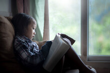 Little Girl Reading Bible On Sofa In Morning At Home. Children's Beliefs Of Christian Concept.
