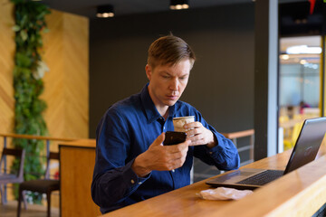 Poster - Young handsome businessman with coffee using phone and sitting with distance at the coffee shop