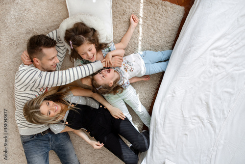 A young beautiful family in casual home look lies on a cozy carpet in the living room. Young dad tickles daughter. The whole family has fun being at home in isolation. Taken from the top view.