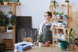 © Seventyfour - Waist up portrait of modern young woman wearing apron standing by wooden table with potted plants and soil ready for potting, home gardening concept, copy space