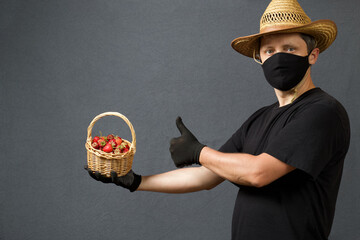 A farmer in a straw hat and a protective mask holds in his hands a wicker basket with strawberries. The second hand shows like. On a gray background. Harvest during the 2020 pandemic.