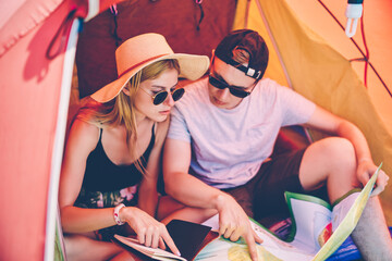 Hipster girl checking travel notes and map for planning hiking route with boyfriend sitting in tent, romantic couple camping during active leisure journey choosing direction for exploring on weekend