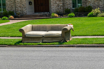 An old torn couch seen on the curb in front of a big house by an asphalt street waiting to be picked up by the trash department