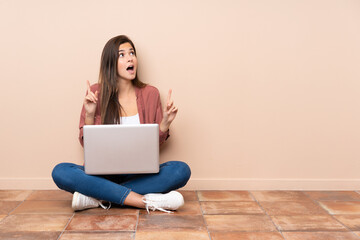 Wall Mural - Teenager student girl sitting on the floor with a laptop surprised and pointing up