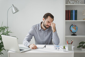 Wall Mural - Exhausted tired young bearded business man in gray shirt sitting at desk work on laptop pc computer in light office on white wall background. Achievement business career concept. Put hand on nose.