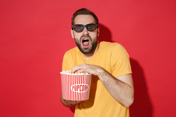 Wall Mural - Shocked scared young bearded man guy in casual yellow t-shirt 3d glasses isolated on red wall background. People emotions in cinema, lifestyle concept. Watching movie film, holding bucket of popcorn.