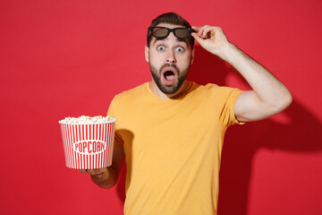 Wall Mural - Shocked young bearded man guy in casual yellow t-shirt 3d glasses isolated on red wall background studio. People emotions in cinema, lifestyle concept. Watching movie film, holding bucket of popcorn.