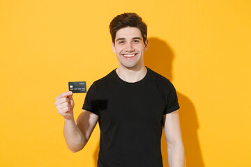 Wall Mural - Smiling young man guy 20s in casual black t-shirt posing isolated on yellow background studio portrait. People sincere emotions lifestyle concept. Mock up copy space. Hold in hand credit bank card.