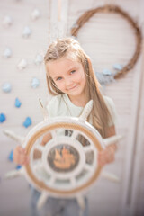 Beautiful little blonde girl holding a white ship steering wheel. At the maritime birthday party. The photo zone is decorated with seashells, stars, a net, lanterns, a rudder, sand. Funny sisters