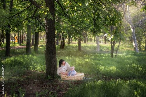 The concept of motherhood and child care. A young mother plays with her little son, who is sitting in a wicker straw cradle. Walk in the park without a stroller sitting on the grass under a chestnut