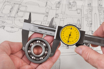 Measurement of steel ball bearing by precise analog caliper above a technical drawing. Metallic measuring tool with round yellow dial in engineer hands. Drafting of combustion engine. Quality control.