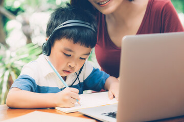 Wall Mural - Young asian mother and son using laptop computer for study and learning together at home, boy writing on notebook for homework and wearing headphone, teacher or mom support child, education concept.