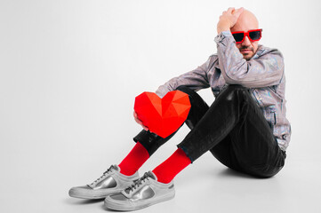 Lovely caucasian man without hair in grey shirt, black leather trousers, red sunglasses and grey trainers shows homemade volume red heart isolated on white background