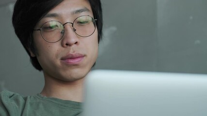 Sticker - A close-up view of a calm young asian man wearing eyeglasses is using his laptop sitting on couch in apartment