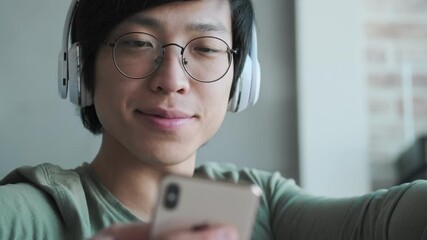 Canvas Print - A happy young asian man with headphones is using his smartphone sitting in the living room at home