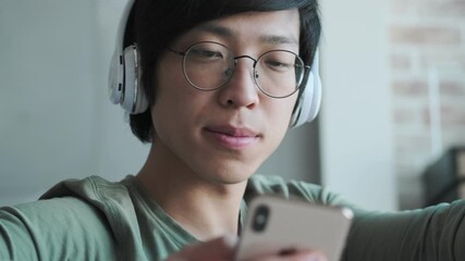 Wall Mural - A close-up view of a handsome young asian man with headphones is using his smartphone sitting in the living room at home