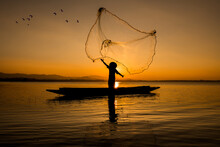 Silhouette Fisherman Throwing Net Casting Fish In Early Morning With Wooden Boat With A Flock Of Birds, Fisherman Life Style