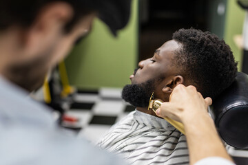 Barber trim hair with clipper on young unshaven black man in barbershop studio. Professional hairdresser cut hair with electric shearer machine on African guy. Male beauty treatment process