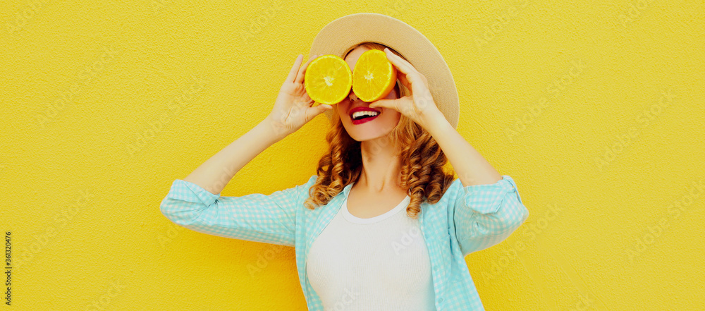 Summer portrait of happy smiling woman covering her eyes with fruits slice of orange wearing straw hat on yellow background - obrazy, fototapety, plakaty 