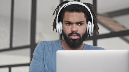 Wall Mural - A serious young african american man wearing headphones is using his silver laptop computer in the living room at home