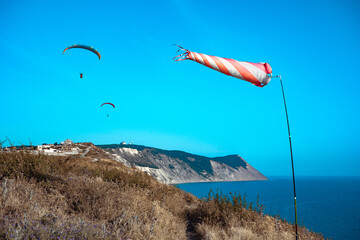 A red and white striped windsock on the seashore amid two flying paragliders. Two paragliders fly against the background of a windsock developing by the wind on the sea coast.