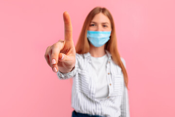 young woman in a medical protective mask on her face, touches the screen with her finger on a pink background
