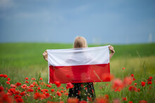 Blond Girl Holding Flag Of Poland In The Poppy Field. Back  View. Polish Flag Day. Independence Day. Travel And Learn Polish Language Concept.