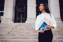 Successful African American Businesswoman Dressed In Trendy White Shirt Standing Near University Building.Charming Student Of Faculty Of Law Holding Folder And Modern Gadgets In Hands And Looking Away