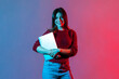 © khosrork - Portrait of successful confident office worker, beautiful girl in casual pullover standing, holding closed laptop or folder, looking at camera with toothy smile. colorful neon light, studio shot