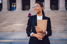 Successful African American Graduate Juridical Specialist Standing Outdoors Against Courthouse Building With Folder In Hands. Prosperous Female Student Of High Economic University In Elegant Wear