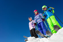 View From Below Of A Group Of Three Children Stand On The Mountain Top In Snow Wearing Ski Colorful Outfit Over Blue Sky