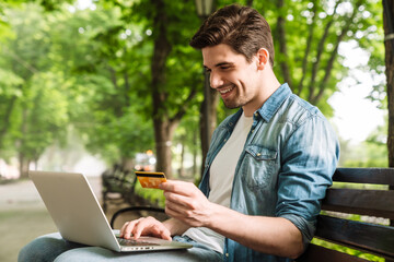 Poster - Photo of man using laptop and holding credit card while sitting on bench