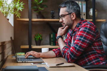 Side view shot of pensive young man sitting at home office and working on laptop
