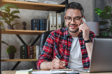 smiling man writing notes while making phone call and using laptop at home