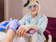 © k8most - manicure master in blue gloves creaming hands of elderly stylish woman in blue sunglasses and jeans jacket sitting at manicure salon (studio) happy. Treatment, care, anti age, manicure concept