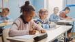 © Gorodenkoff - In Elementary School Classroom Brilliant Black Girl Writes in Exercise Notebook, Taking Test and Writing Exam. Junior Classroom with Diverse Group of Children Working Diligently and Learning New Stuff