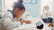 © Gorodenkoff - In Elementary School Classroom Brilliant Black Girl Writes in Exercise Notebook, Taking Test. Junior Classroom with Diverse Group of Bright Children Working Diligently and Learning. Side View Portrait