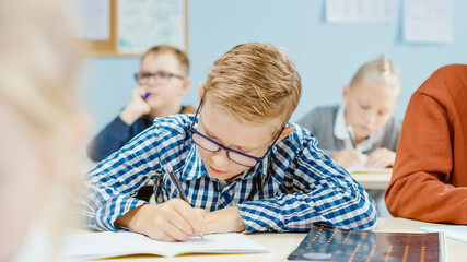 Wall Mural - In Elementary School Class: Portrait of a Brilliant Caucasian Boy Wearing Glasses Writes in the Exercise Notebook. Diverse Group of Bright Children Learning Science and Creative Thinking