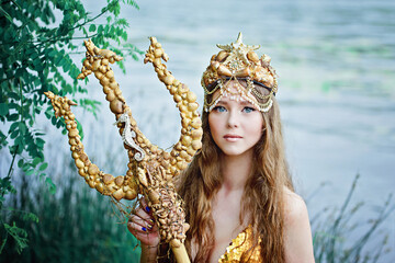 Fantasy woman real mermaid with trident myth goddess of sea with golden tail sitting in sunset on rocks.. Gold hair crown shells pearls jewelry. Mermaid sitting on shore. fantasy concept.