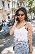 Portrait of a young beautiful woman having mobile phone conversation while standing on the street in sunny summer day, attractive female talking on cell telephone during walking in urban setting