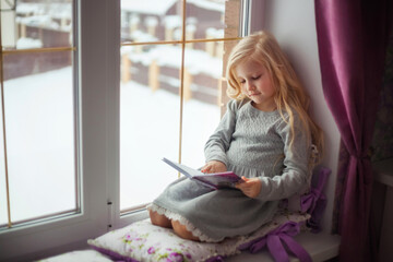 A cozy portrait of a cute child of a blonde girl in a knitted dress lying on the window in the children's room and reading a book. Pastime in the fall or winter. Children read books, home schooling