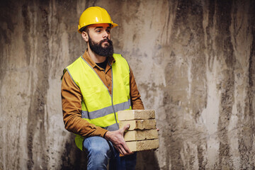 Wall Mural - Young bearded worker in working clothes standing at construction site and holding bricks.