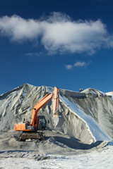 Wall Mural - Red excavator stands next to a hill of crushed stone on the background of a blue sky with clouds, close-up.