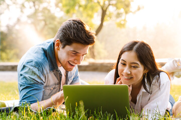 Wall Mural - Image of couple using laptop and smiling while lying on grass