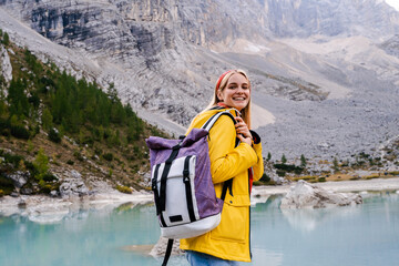 Wall Mural - Lake Sorapis. Dolomites Alps. Italy. Blonde girl in yellow raincoat with backpack stays in the middle of azure water & smiles on background of pine forest & rock mountains